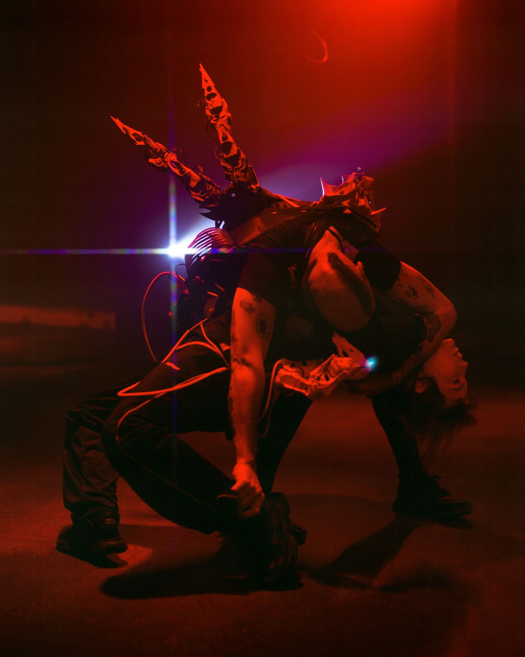 A person with Iro-haircut, kneeling on the floor. Their upper body faces the floor. They were cloth with big "goads". The scene is in red light.