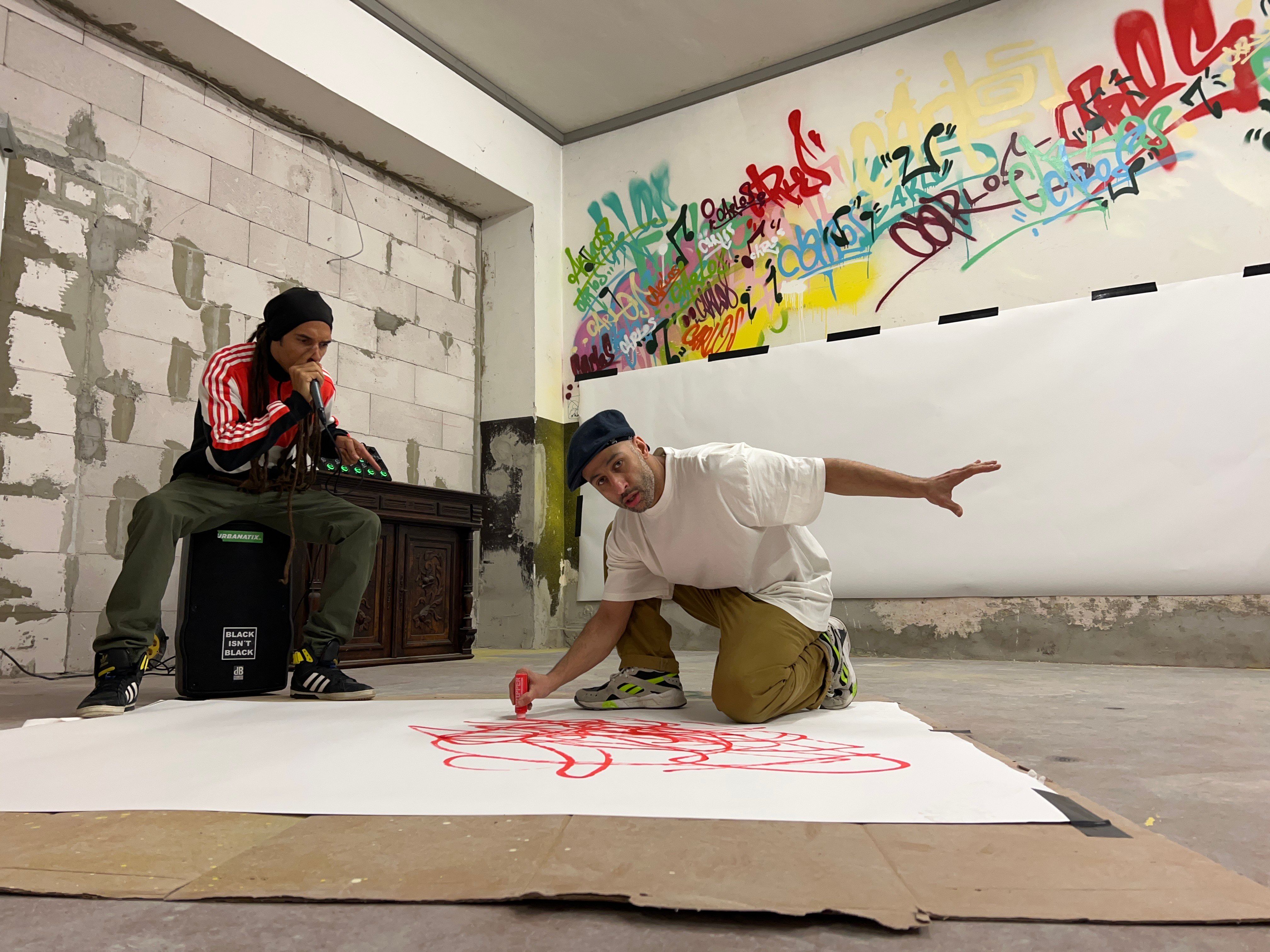 Two people can be seen in the photo. One person is sitting on a jukebox and speaking into a microphone. The other person is kneeling on the floor and drawing something on paper with a red marker and  is looking into the camera.