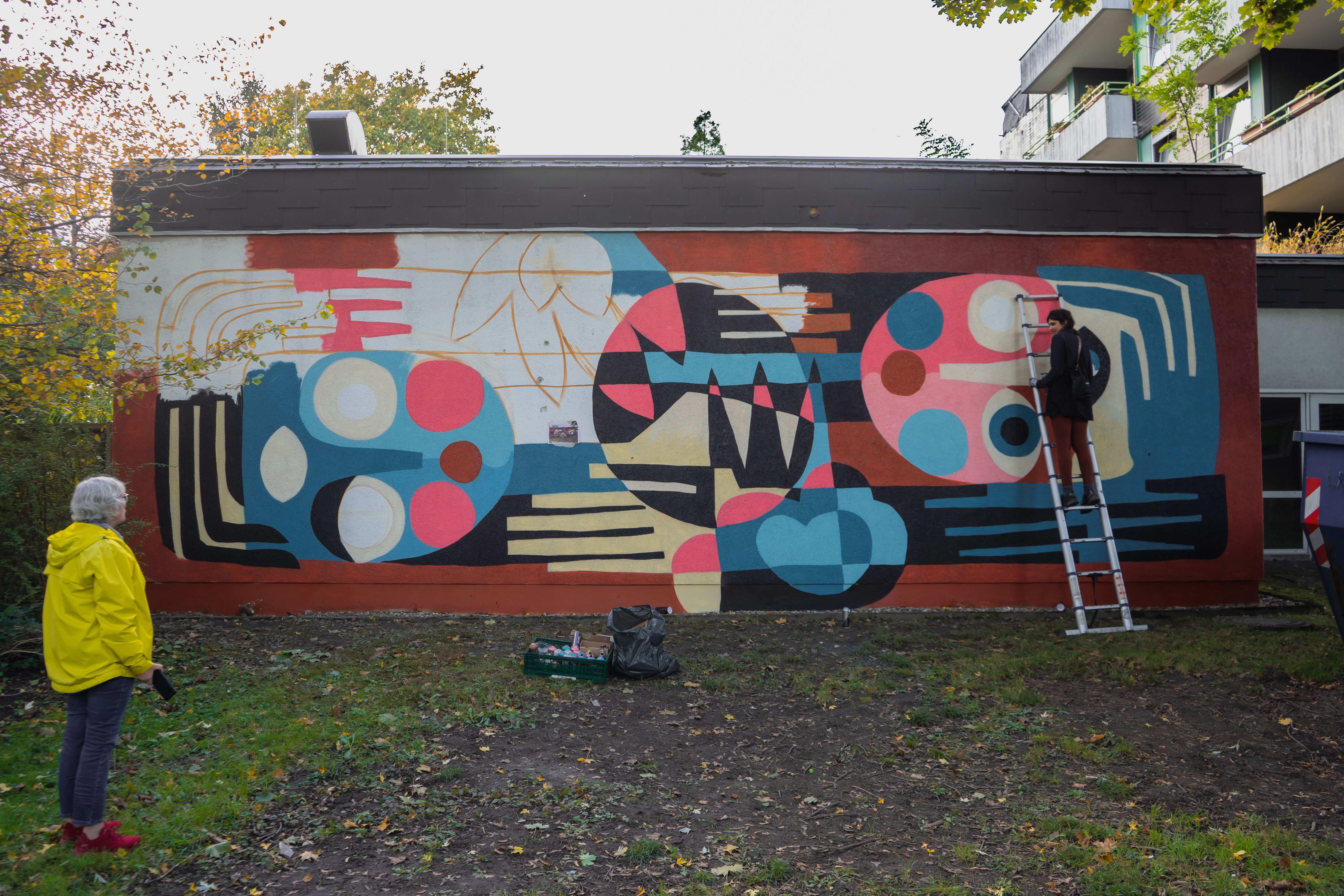 A painted house wall can be seen. A person is standing on a ladder leaning against the wall. On the left of the picture is another person looking at the wall.