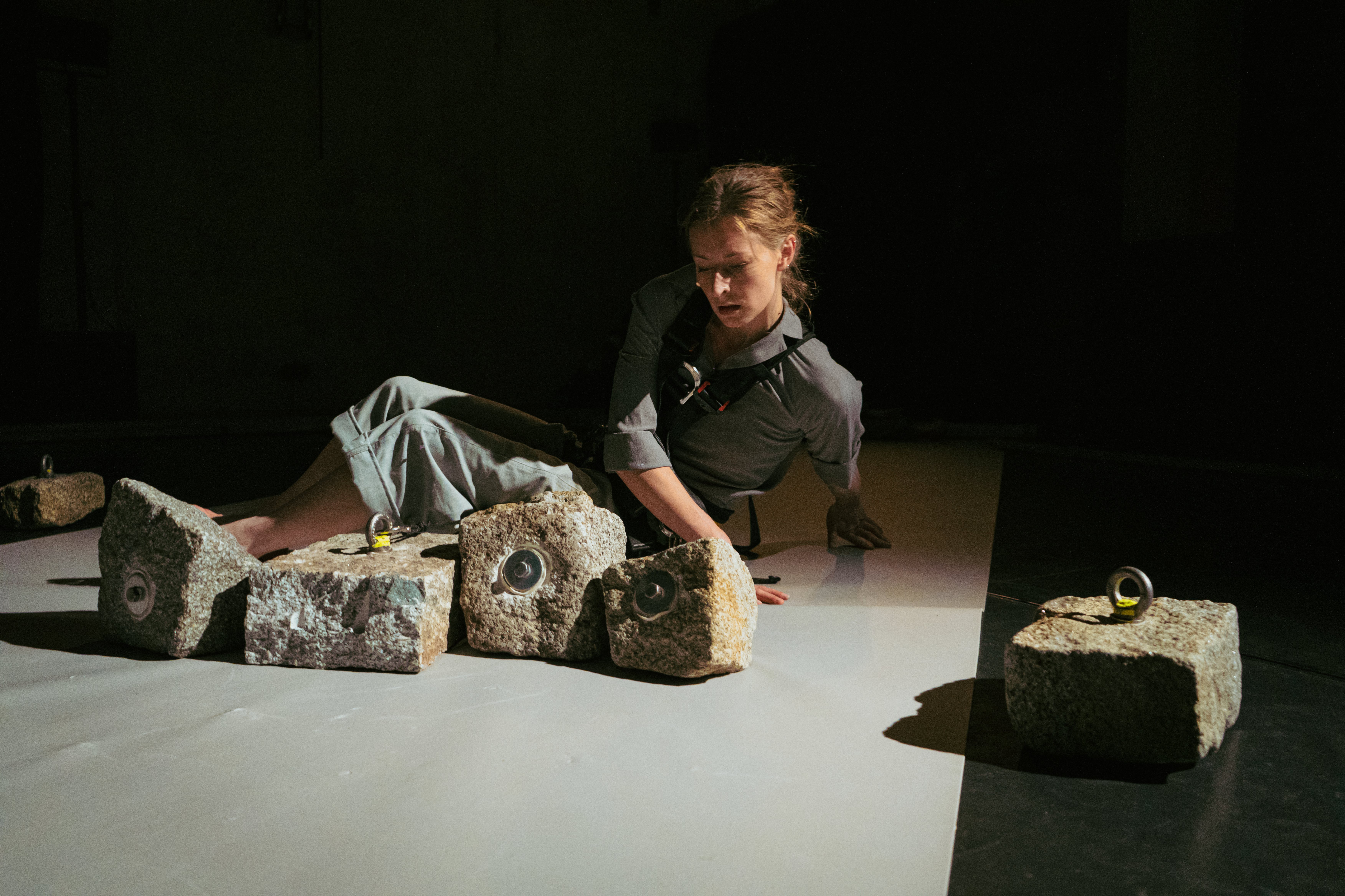 A person sits on the floor and rests their hands on the ground at their sides. Large stones lie in front of the person. Metal hooks are attached to the stones.