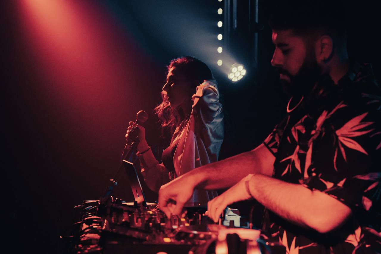 A musician playing the guitar can be seen out of focus in the foreground. The focus of the picture is on several people dancing in front of a building, looking in the direction of the musician.