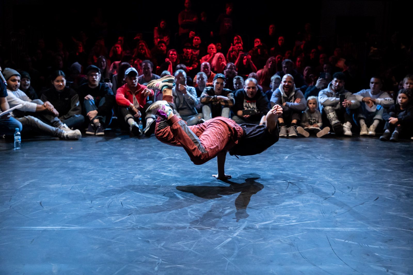 You can see a person breakdancing and leaning on one arm off the ground. An audience sits in front of the person and watches them.