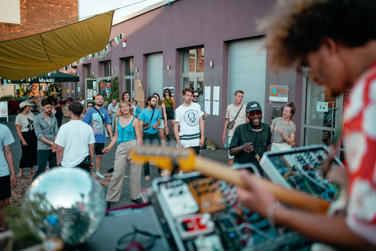 A musician playing the guitar can be seen out of focus in the foreground. The focus of the picture is on several people dancing in front of a building, looking in the direction of the musician.
