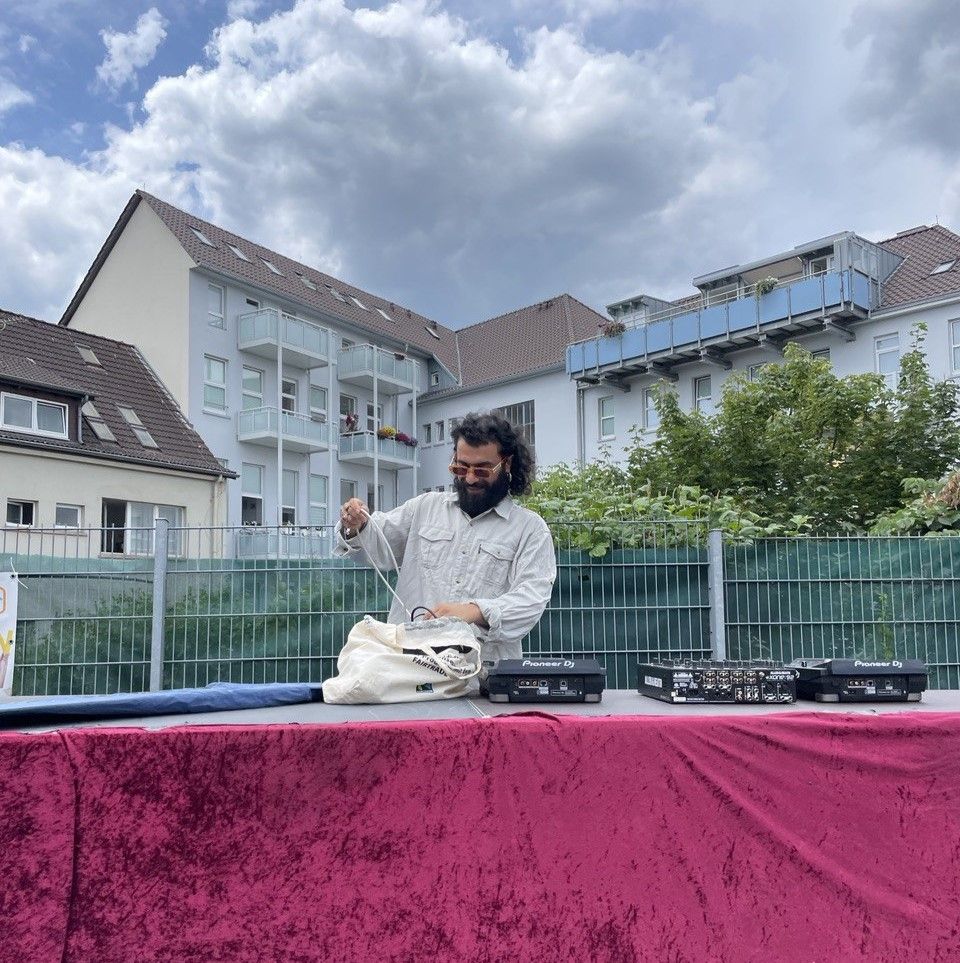A person sets up a DJ deck outside on a table with red velvet. In the background a fence and houses.