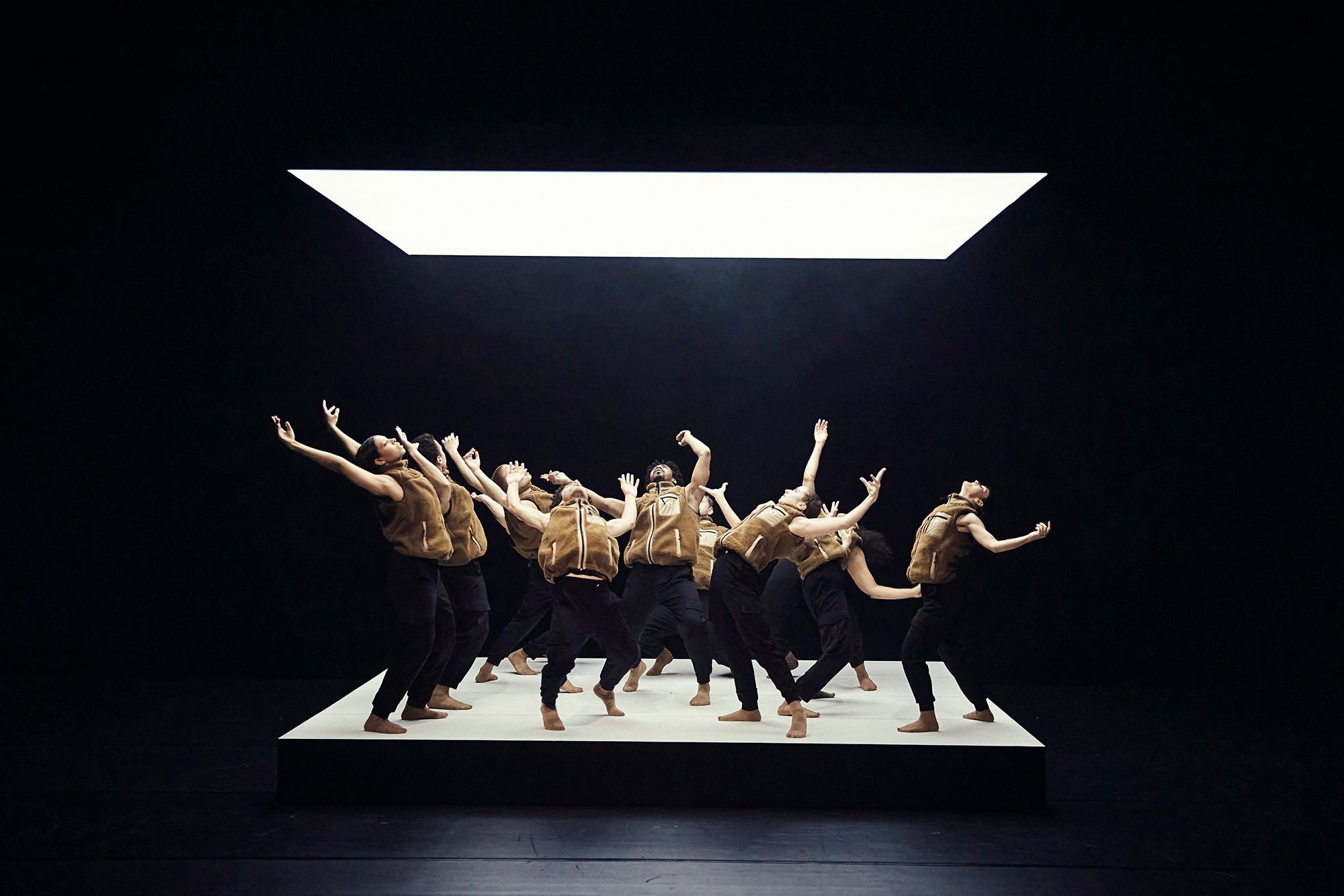 The photo shows the dancers in of the production “Same Love”. They are standing on a white square platform. They are looking into the square light source hanging above them and have their arms outstretched. Their bodies are bent backwards.