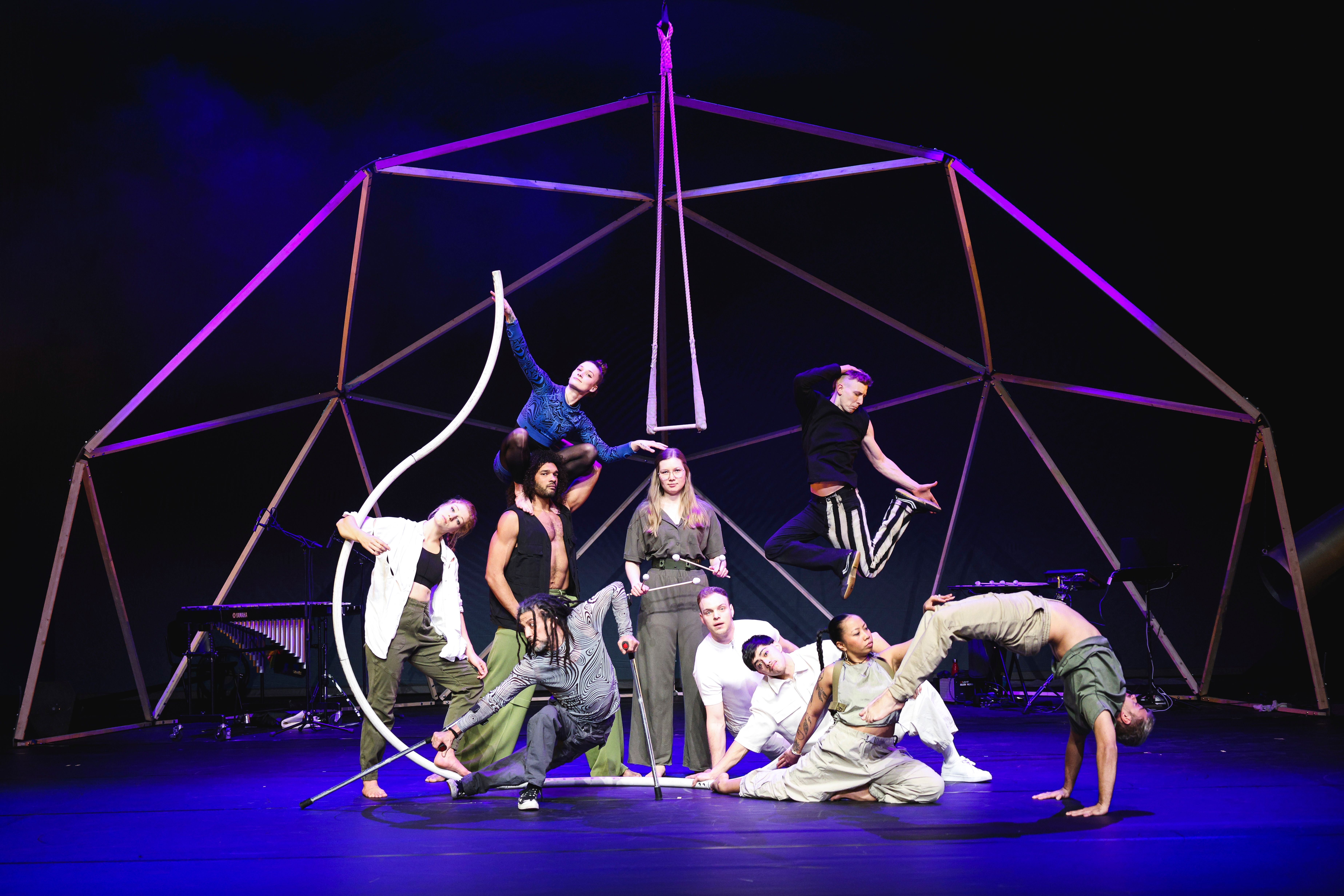 A group of acrobats stand and sit on the stage. They have all assumed different poses and are looking into the camera.