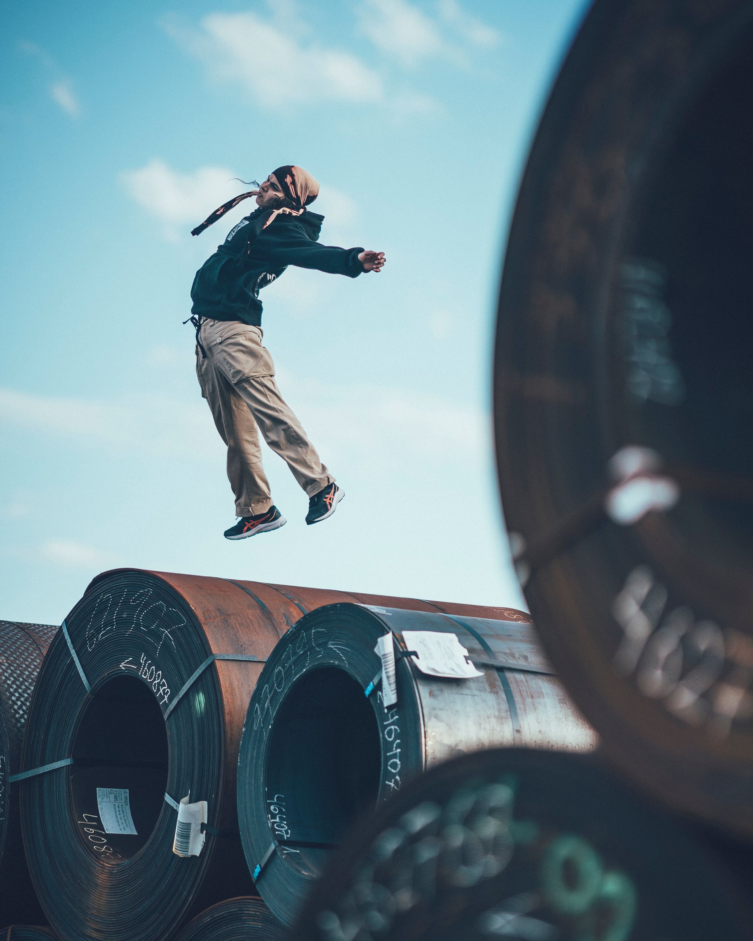 A dancer jumps into the air and spreads her arms and legs. Underneath her are large steel pipes. She is probably on an industrial site.