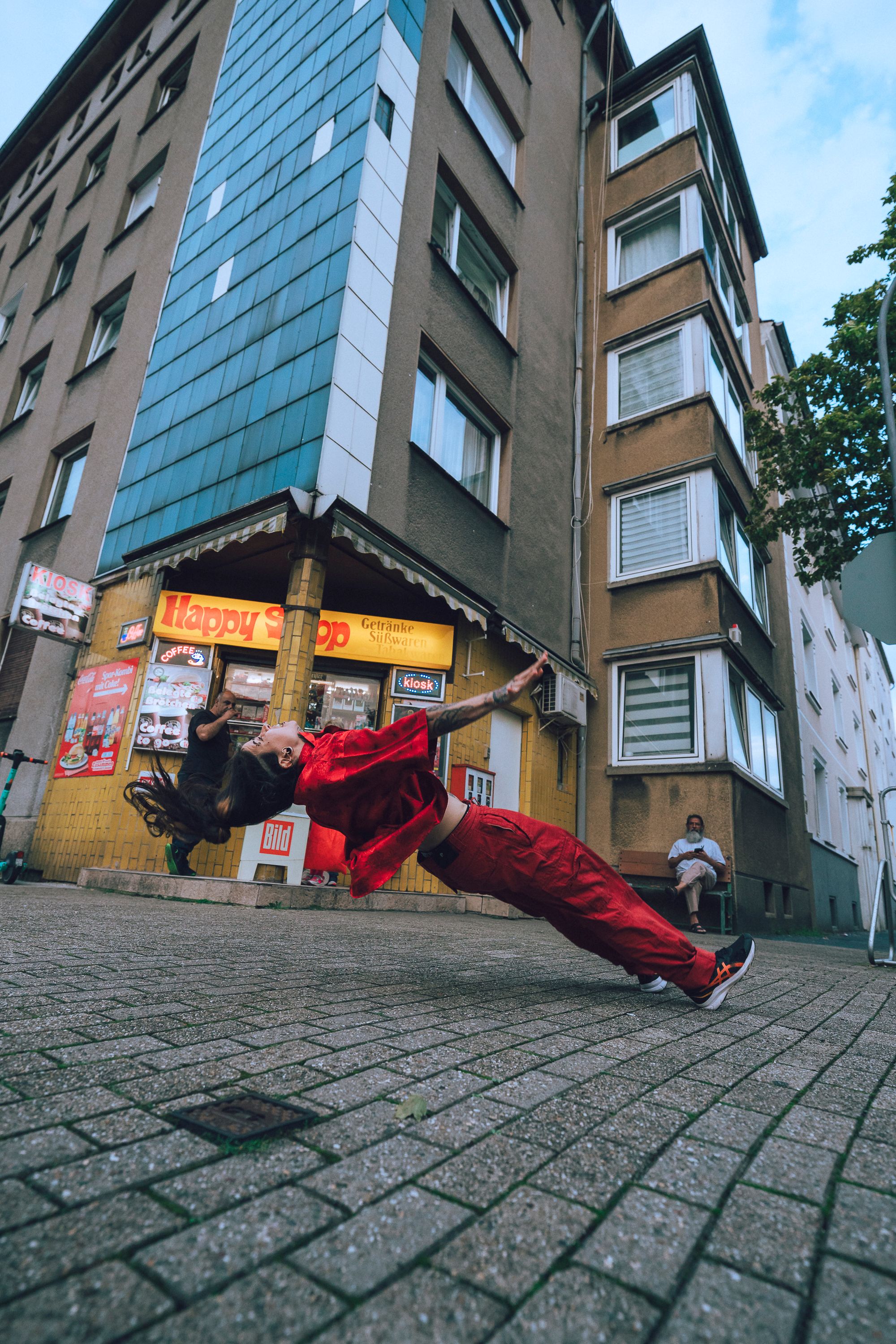 There is a dancer dressed in red, in front of a shop. She is leaning back with her body.