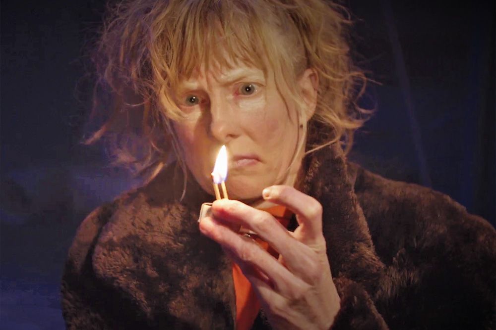 You can see a person with blonde hair. The person is holding a lit match in their hand and is staring at the flame.
