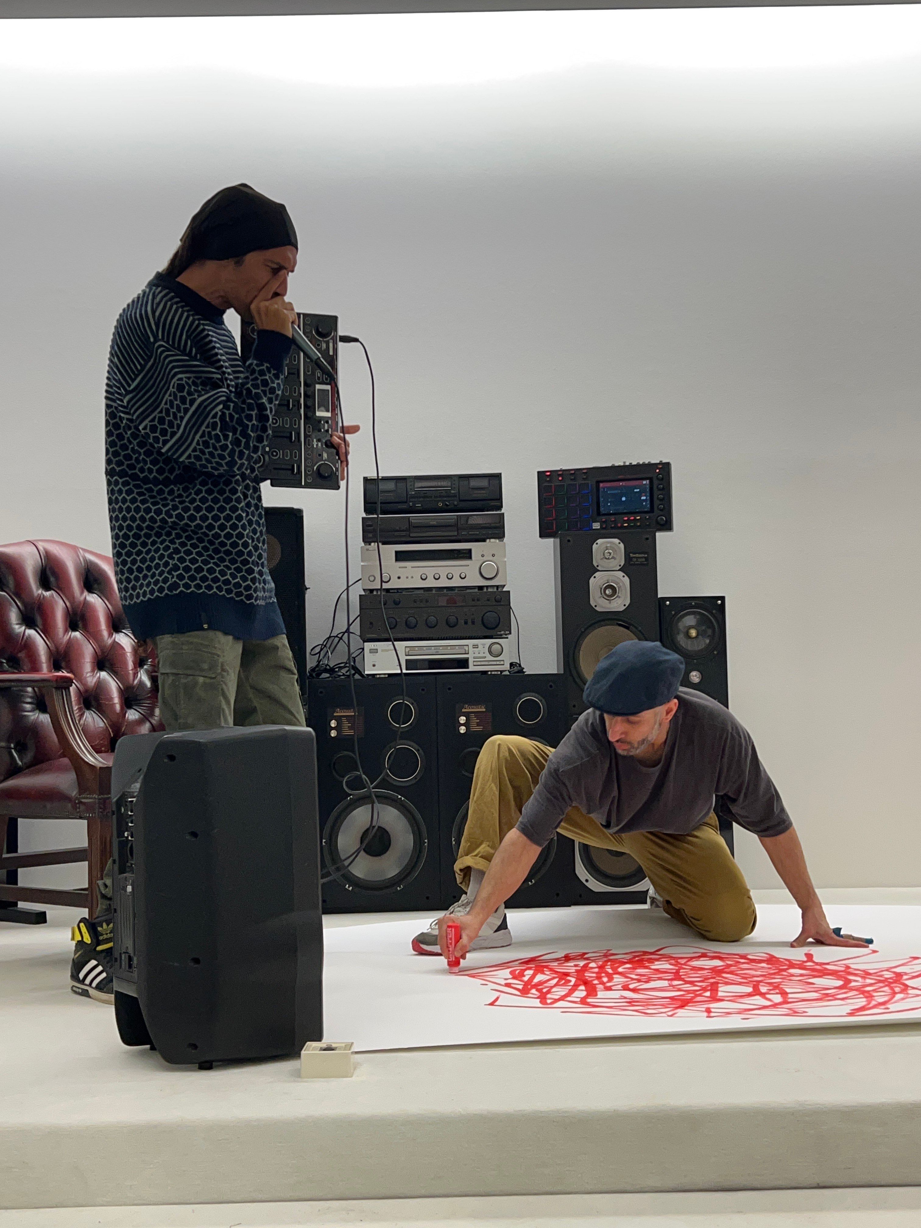 Two people can be seen in the picture. One person is standing and holding a microphone. The person is speaking into the microphone. The other person kneels on the floor and draws something on paper with a red marker.