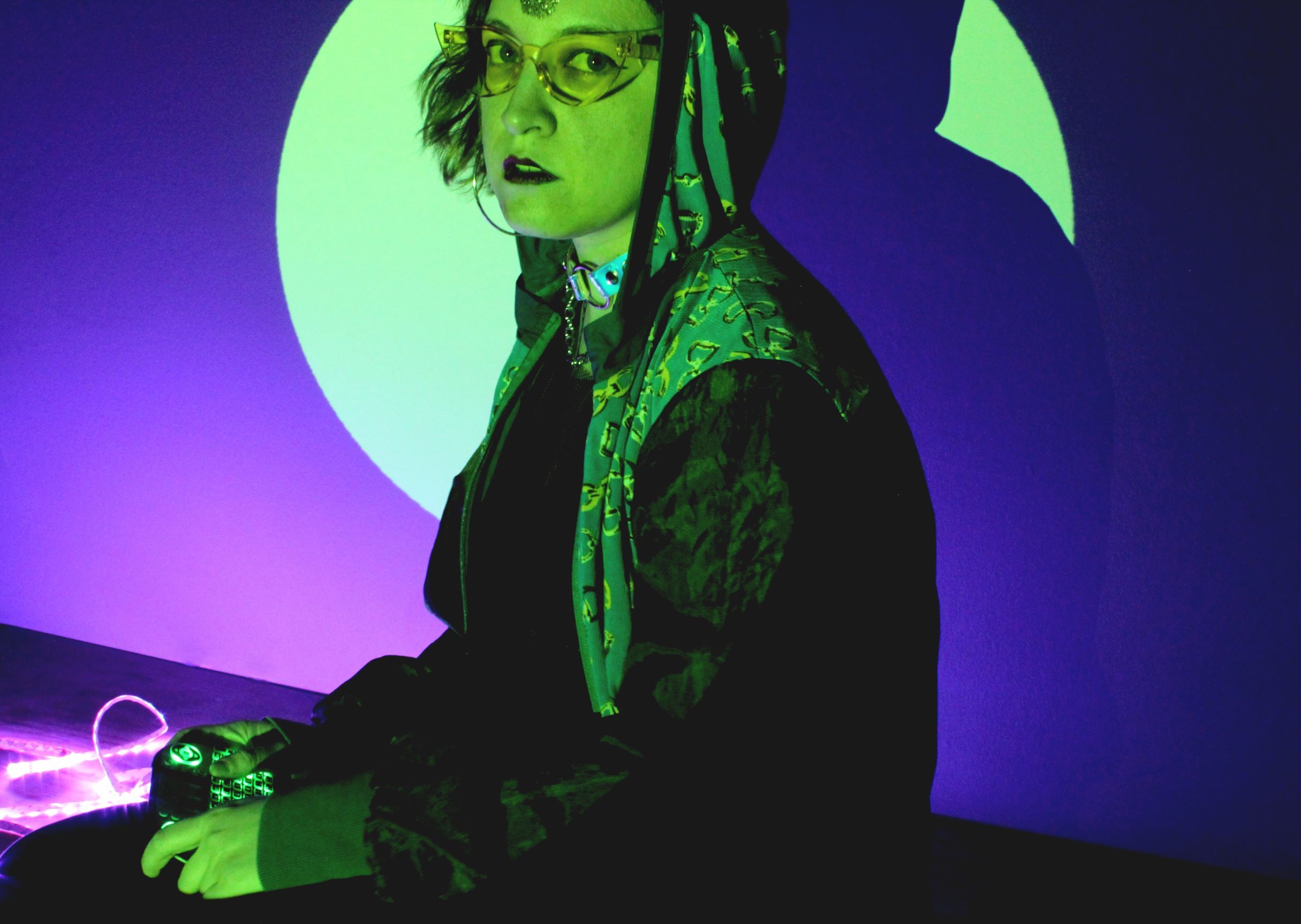 The upper body of a person wearing glasses with shoulder long hair. They are seated in green light and wear a hoodie. The background is violett and blue.