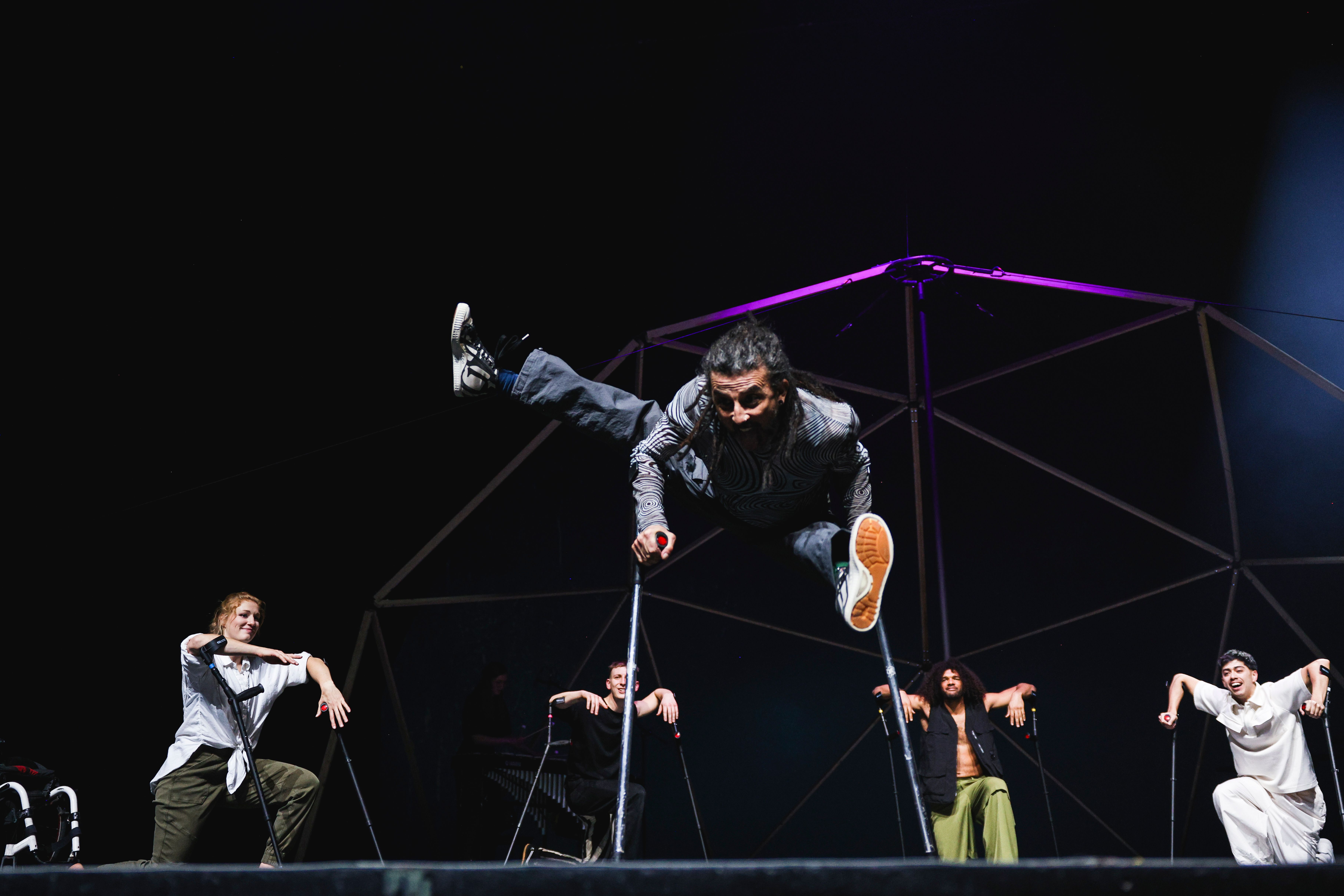 A person with crutches can be seen in the foreground of the picture. The person is performing acrobatics with the help of the crutches. A group of people can be seen in the background. They are watching the person in the foreground during the performance.