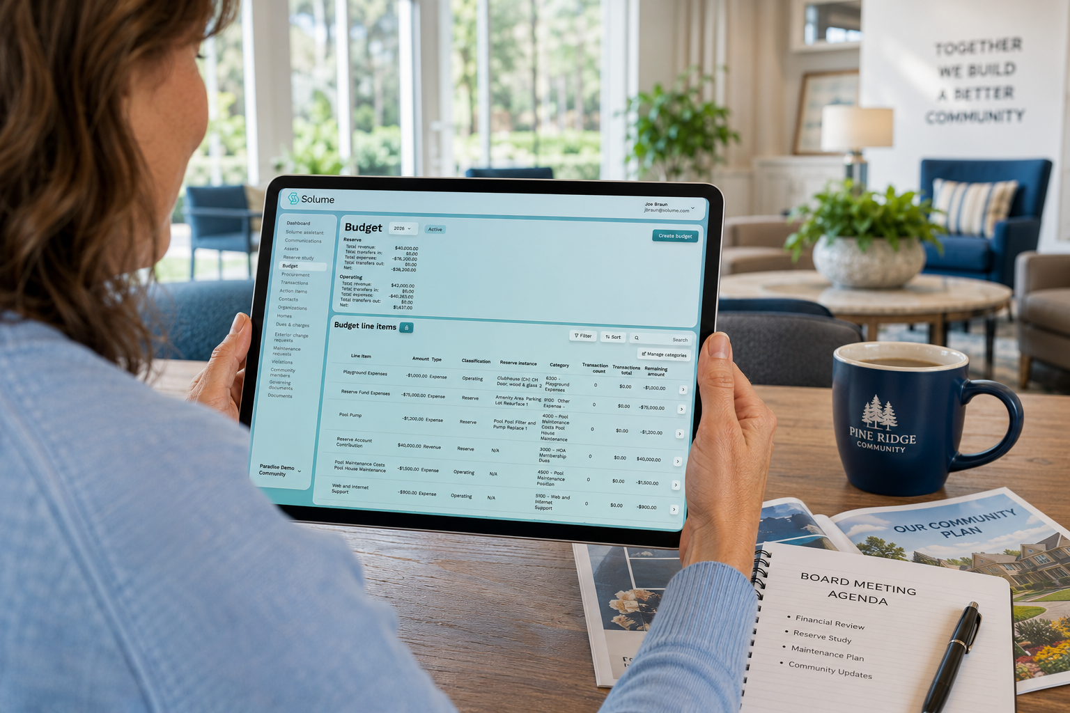 Volunteer HOA board member reviewing the Solume budget management dashboard on a tablet in a bright community clubhouse, showing reserve and operating budgets, budget line items, and financial planning tools.
