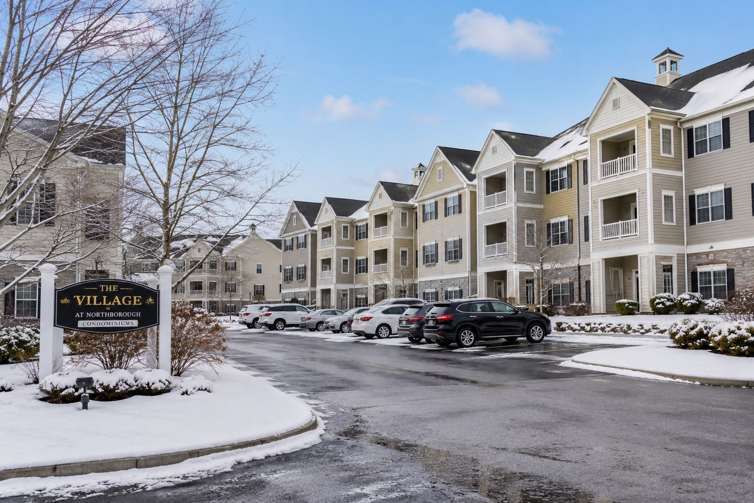 Wide-angle view of a well-maintained Massachusetts condominium complex during winter, featuring multiple multi-story residential buildings with snow-covered grounds, clear parking areas, intact roofs, parked vehicles, and bare trees under natural daylight.