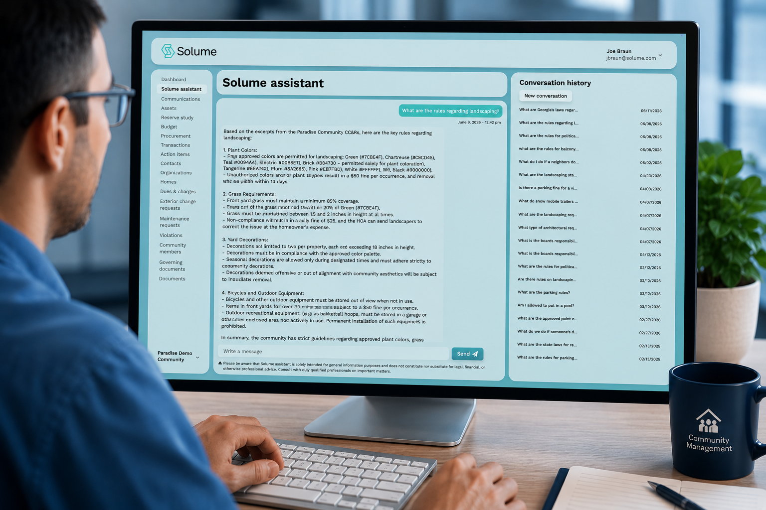 Over-the-shoulder view of a desktop monitor displaying the Solume Assistant dashboard with HOA community management features, AI-powered conversation history, and community governance information in a modern office workspace.