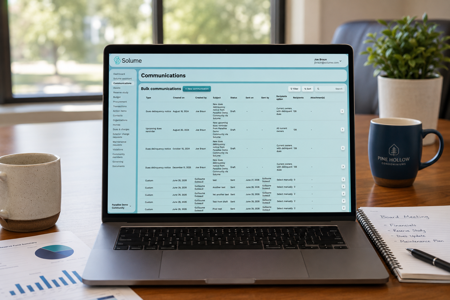 A laptop on a wooden conference table displays the Solume communications dashboard, showing bulk homeowner communications, dues reminders, and delinquency notices. The workspace includes coffee mugs, a notepad, and financial reports, with soft natural daylight creating a clean, professional office environment.