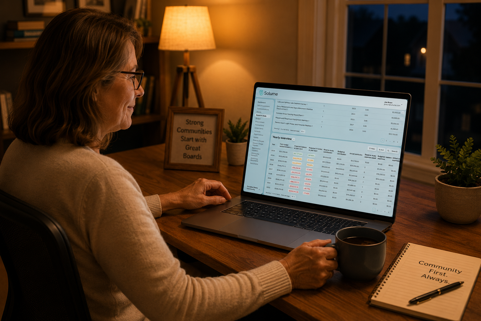 Volunteer board member working at a home office desk in the evening, viewing the Solume Reserve Study dashboard on a laptop while holding a coffee mug in a warmly lit room.