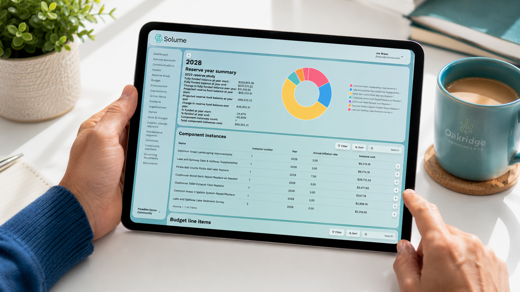 Close-up of a volunteer HOA board member holding a tablet displaying the Solume reserve study dashboard with financial charts, reserve summaries, and budget line items on a clean, modern desk in bright natural light.