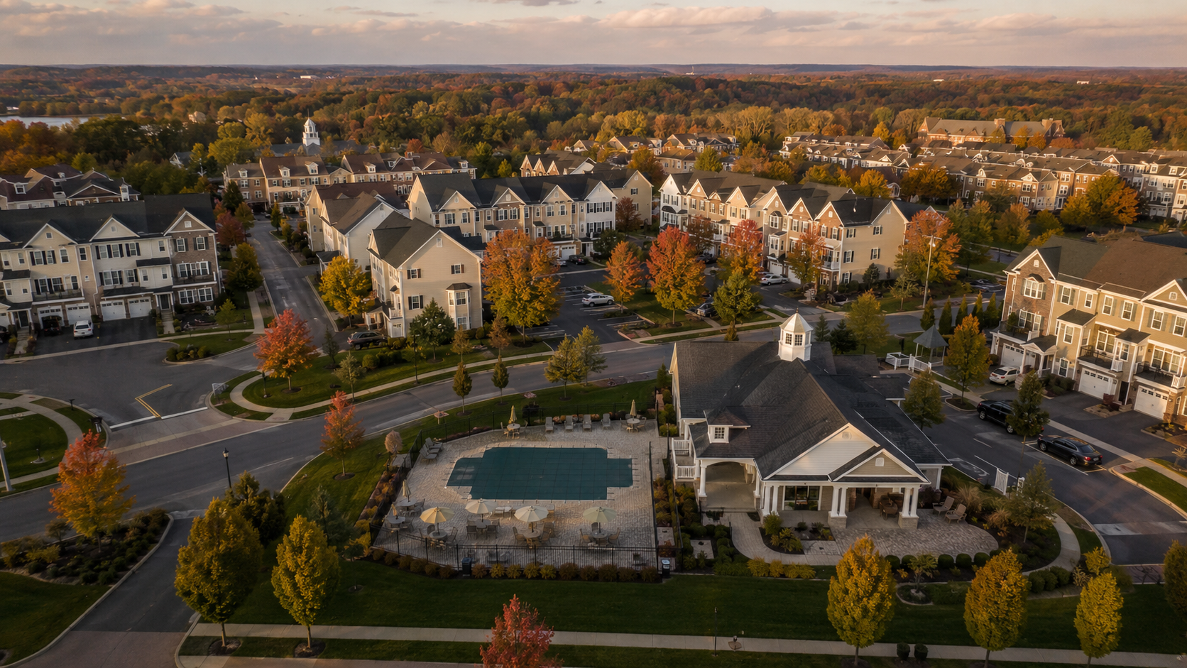 Aerial view of a well-maintained New Jersey townhome community featuring tree-lined streets, shared roads, a central clubhouse, and vibrant autumn foliage under warm late-afternoon golden light.