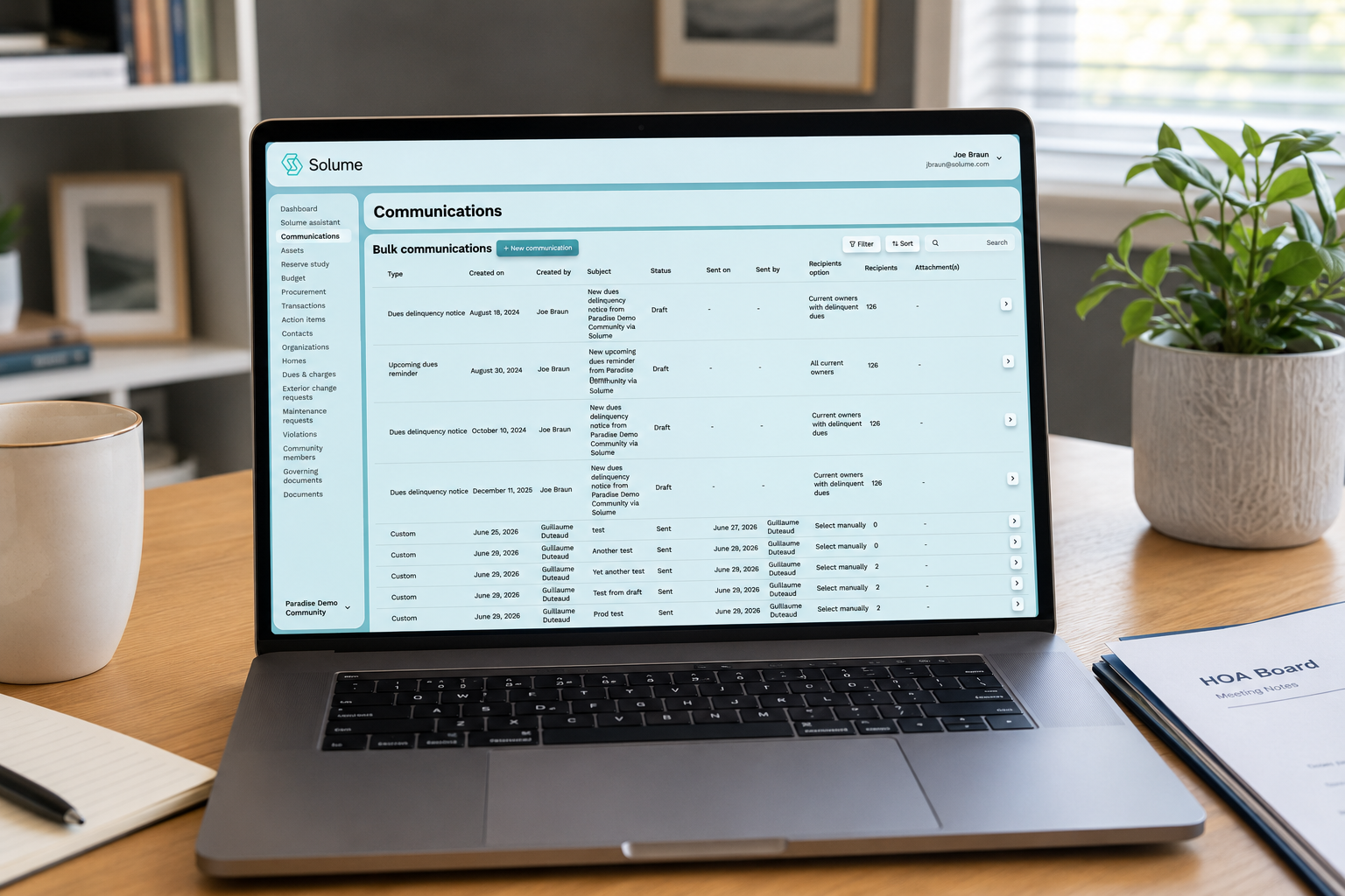 Laptop on a home office desk displaying the Solume HOA communications dashboard with bulk communication records, filters, recipient lists, and management tools, alongside a notebook, coffee mug, and potted plant.