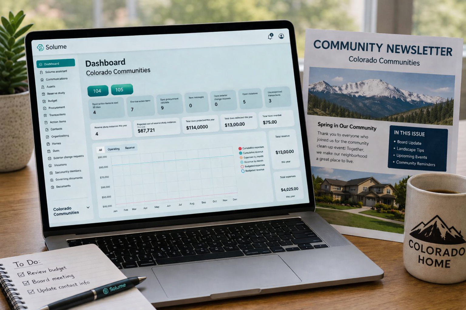 Laptop displaying the Solume HOA management dashboard beside a Colorado community newsletter on a wooden desk in a naturally lit home office setting.