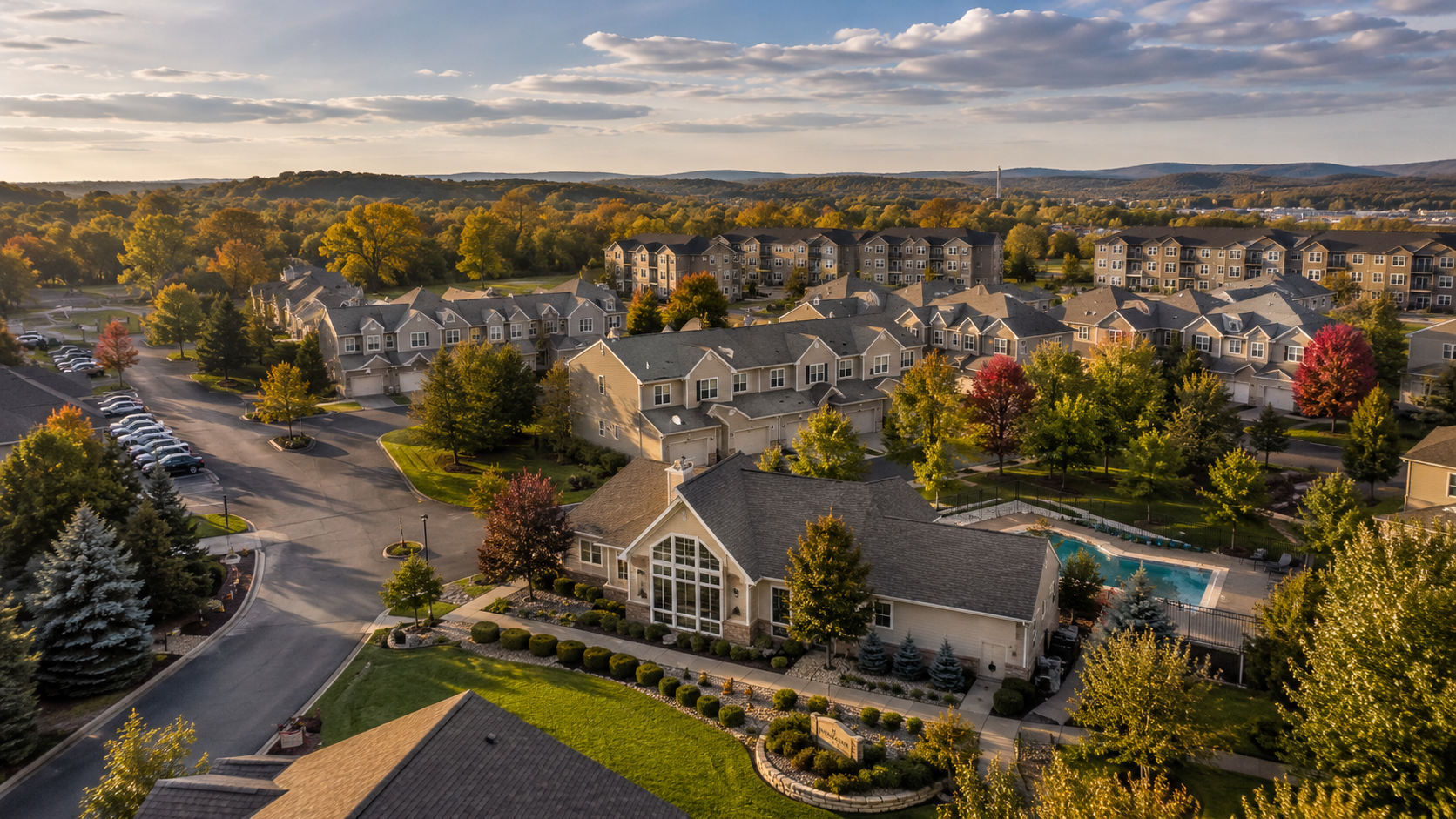Aerial view of a well-maintained Missouri townhome and condominium community during early autumn, featuring tree-lined streets, a central clubhouse, clean roads, and warm golden-hour lighting beneath a partly cloudy sky.