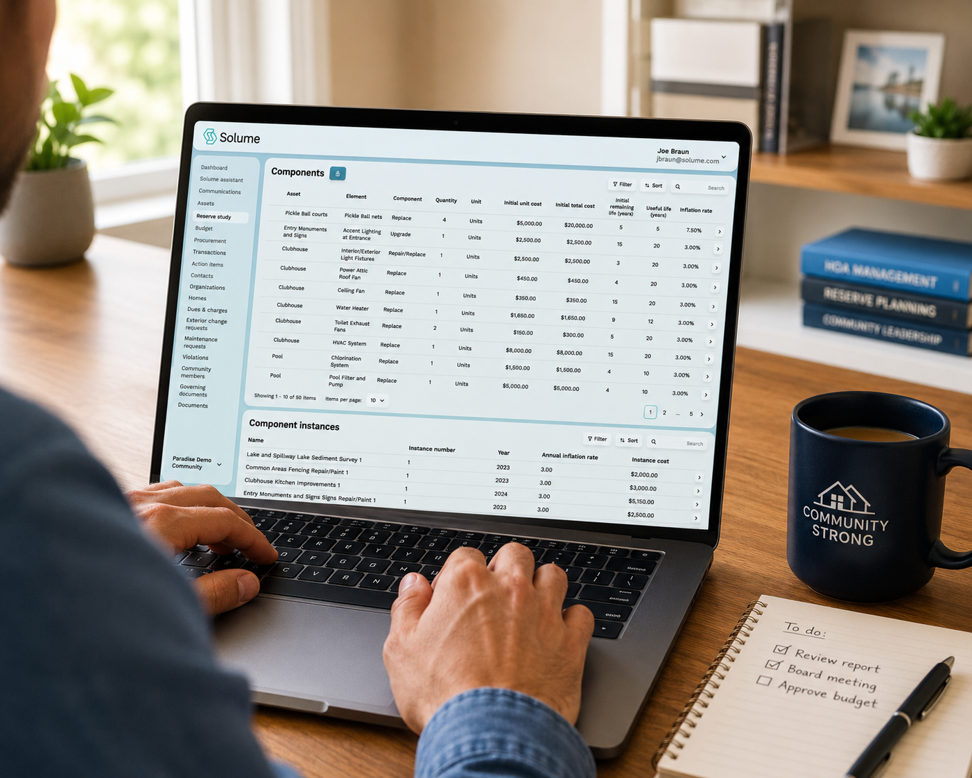 Over-the-shoulder view of a person using a laptop displaying the Solume reserve study dashboard with community asset components, replacement schedules, and cost data, on a tidy wooden home office desk with a coffee mug and notepad in soft morning light.