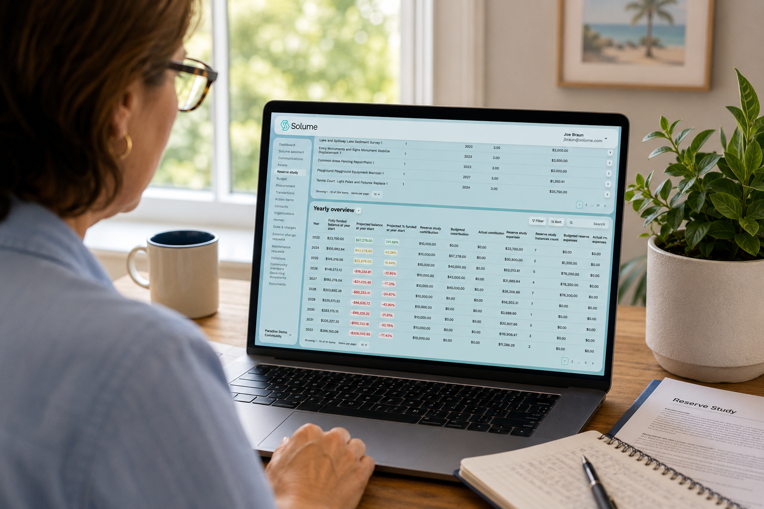 Over-the-shoulder view of a board treasurer working on a laptop displaying the Solume Reserve Study dashboard with funding projections, yearly reserve overview, and financial planning data in a bright home office.