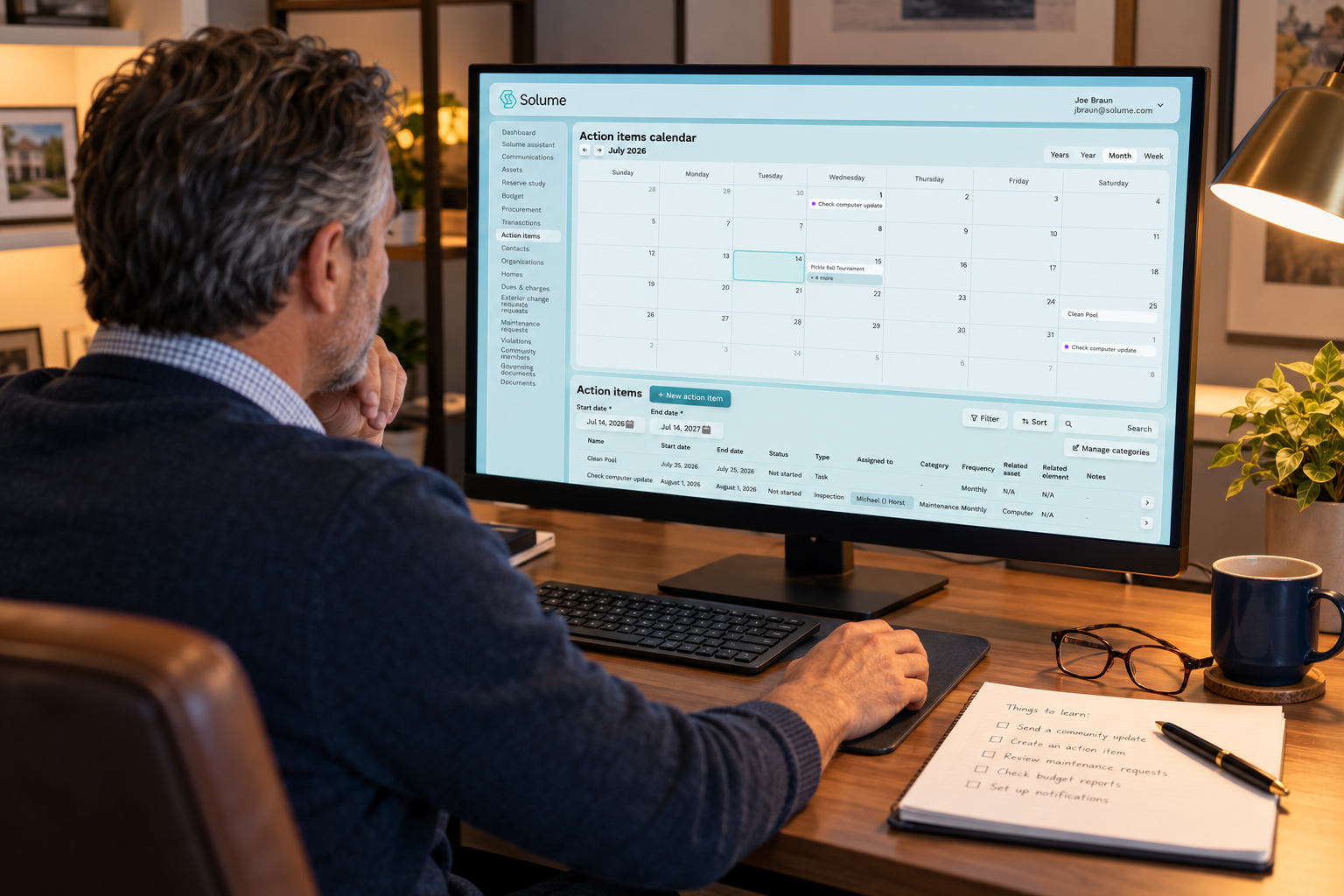 Middle-aged volunteer board member learning to use HOA management software on a desktop monitor in a warmly lit home office, with a notepad, reading glasses, and coffee on the desk.