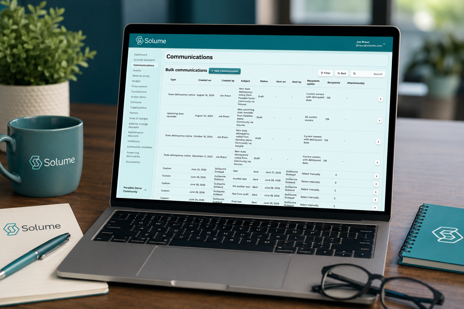 Laptop displaying the Solume communications dashboard with bulk communication records, message statuses, and resident recipient lists on a clean office desk with branded stationery.