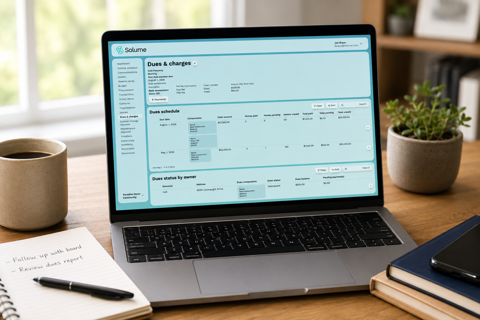 A modern laptop on a clean home office desk displays the Solume HOA "Dues & Charges" dashboard, with payment schedules, owner dues status, and financial details visible alongside a coffee mug and notepad.