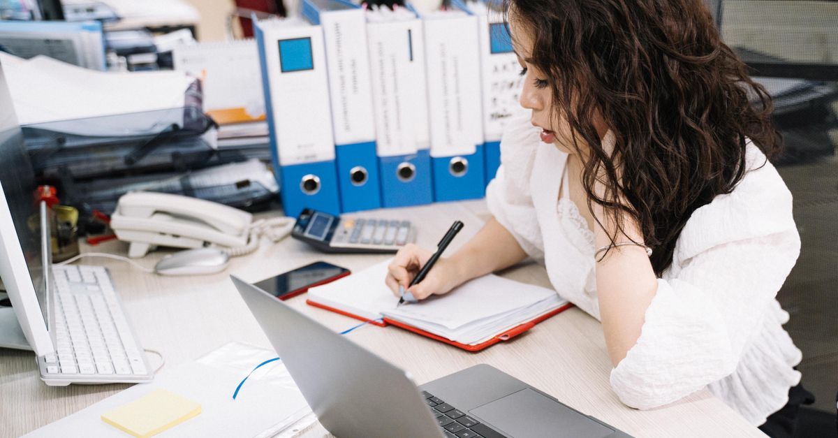A person takes handwritten notes while using a laptop and paper documents.
