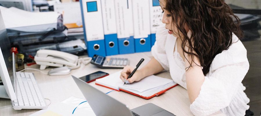 A person takes handwritten notes while using a laptop and paper documents.