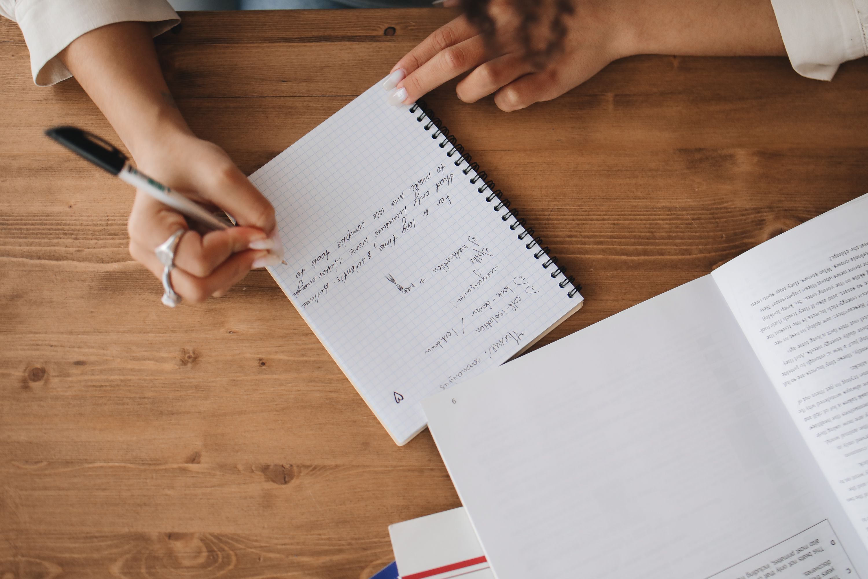 Hands taking notes in a spiral notebook beside an open book.