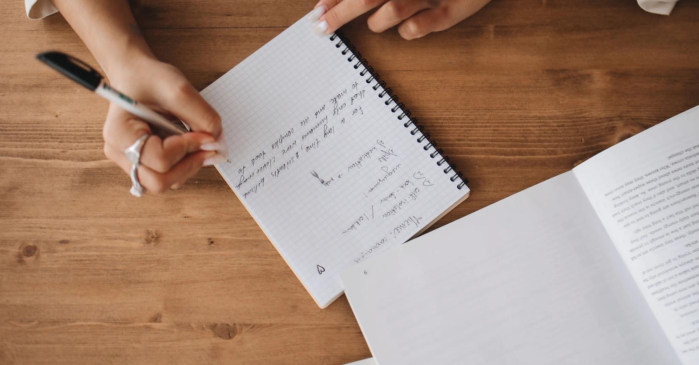 Hands taking notes in a spiral notebook beside an open book.