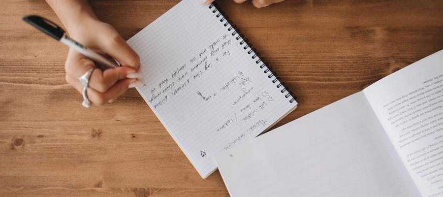 Hands taking notes in a spiral notebook beside an open book.