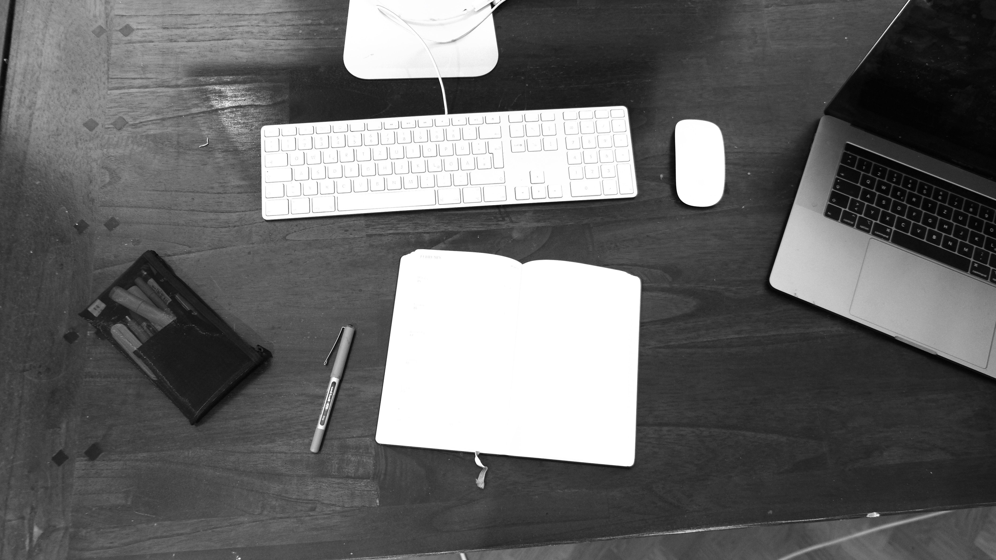 Black-and-white view of a notebook, keyboard and dark-screen laptop on a desk.