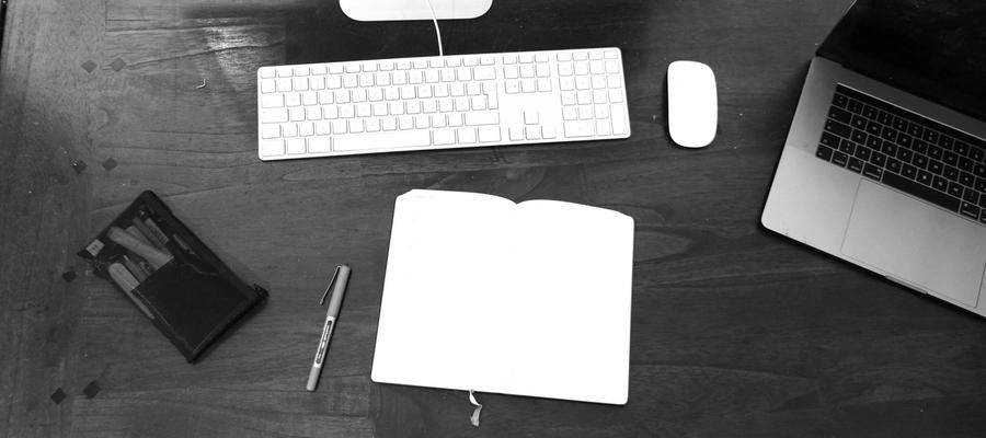Black-and-white view of a notebook, keyboard and dark-screen laptop on a desk.