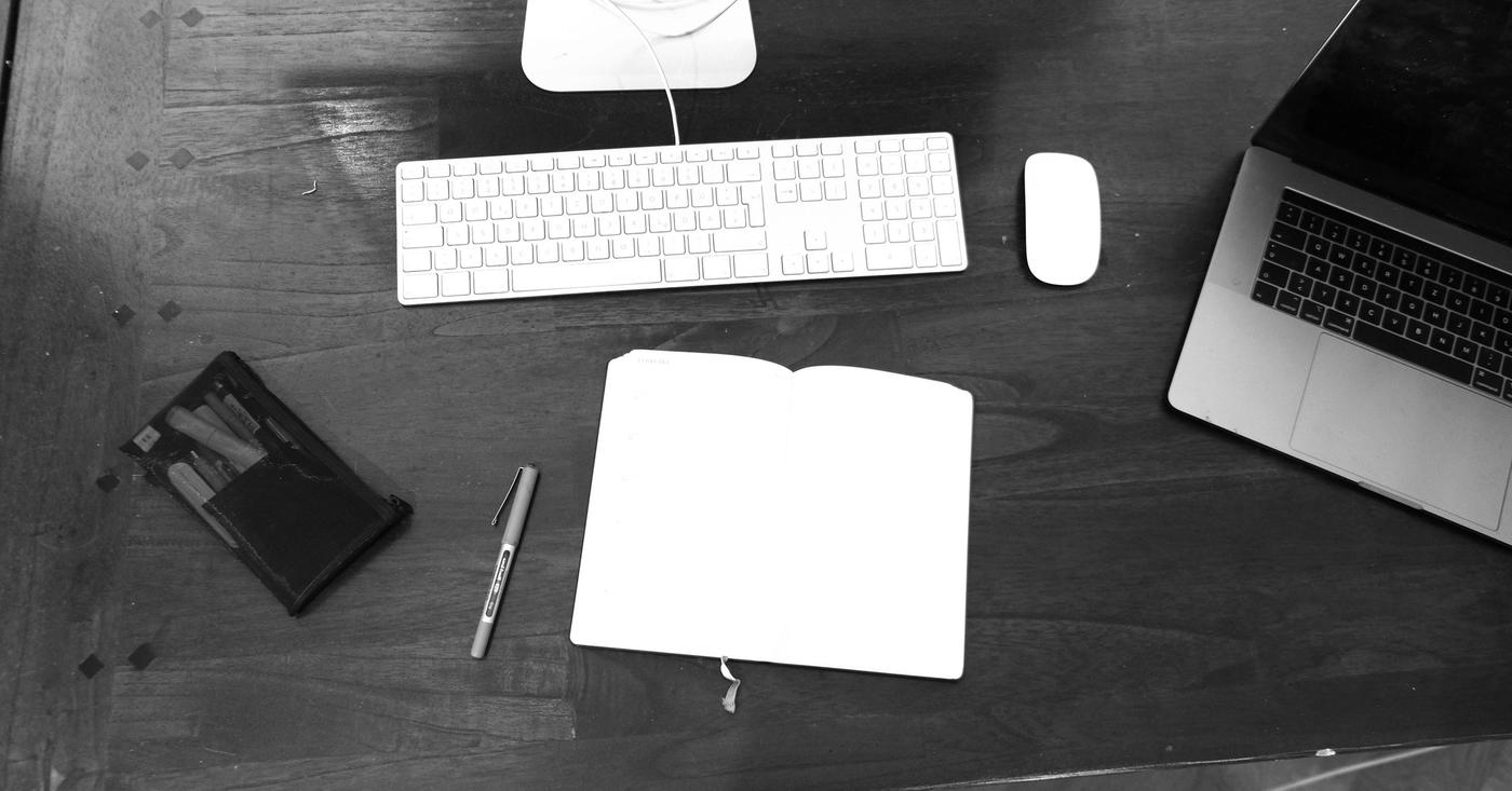 Black-and-white view of a notebook, keyboard and dark-screen laptop on a desk.