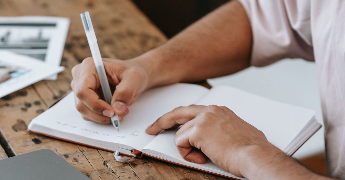 A person takes notes in a notebook beside a laptop.