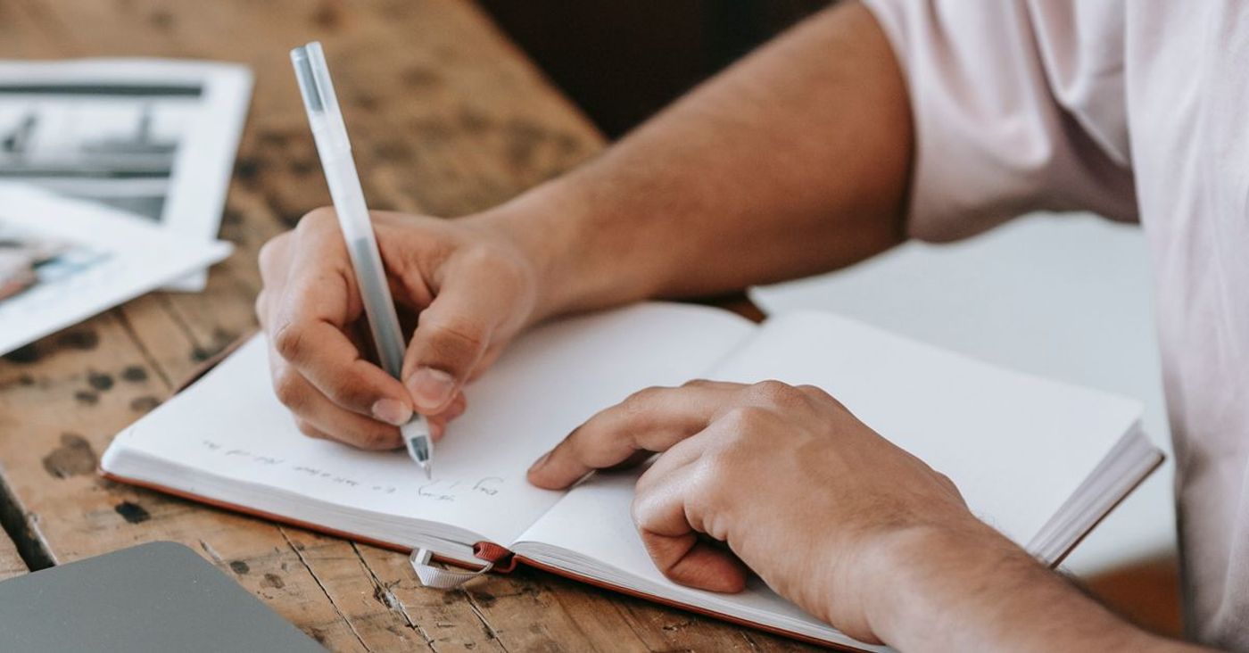 A person takes notes in a notebook beside a laptop.