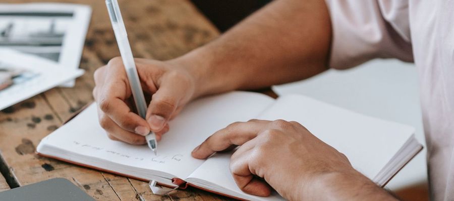 A person takes notes in a notebook beside a laptop.