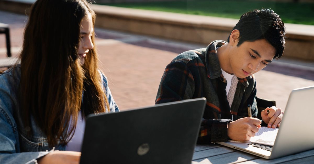 Two students use laptops and take notes at an outdoor table.