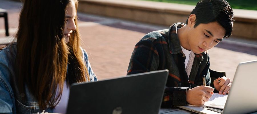 Two students use laptops and take notes at an outdoor table.