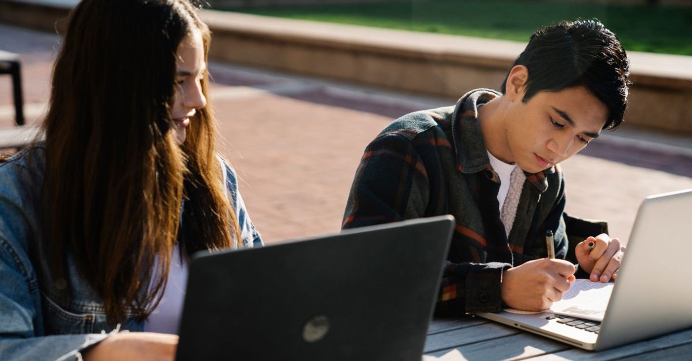Two students use laptops and take notes at an outdoor table.