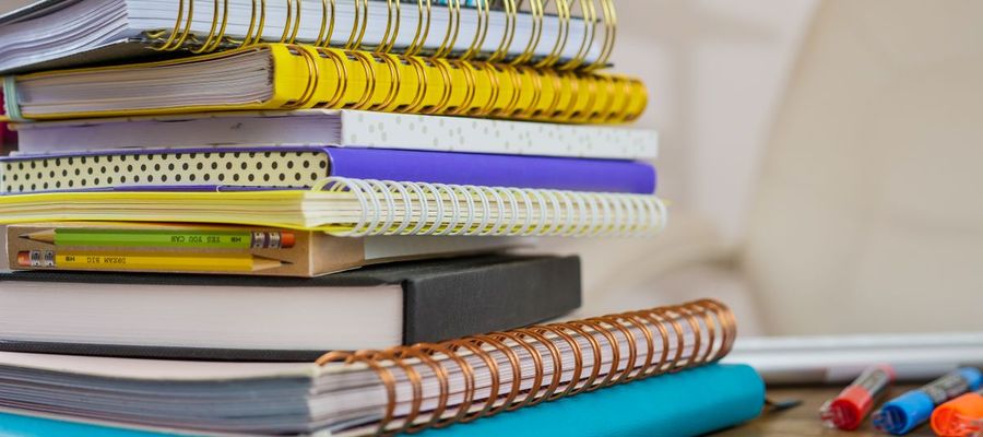 A stack of colourful spiral notebooks and pencils on a wooden desk.