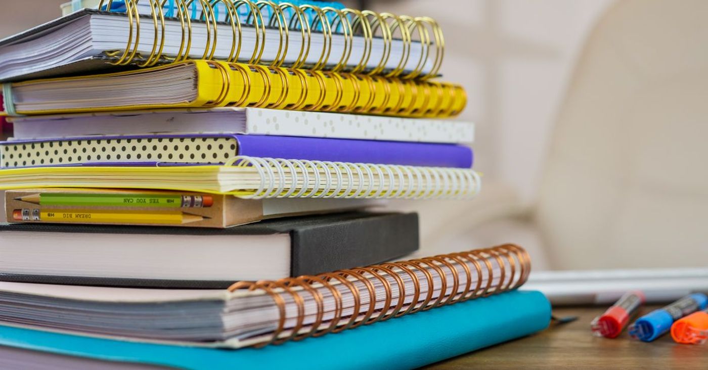 A stack of colourful spiral notebooks and pencils on a wooden desk.