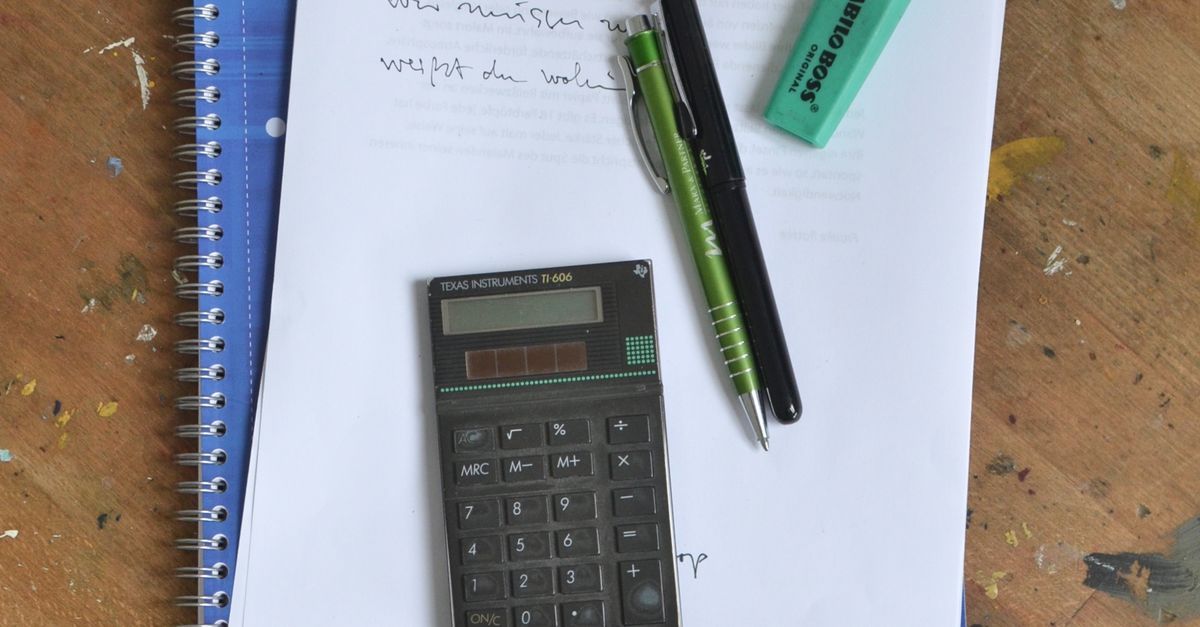 A notebook, paper notes, calculator and pens on a wooden desk.