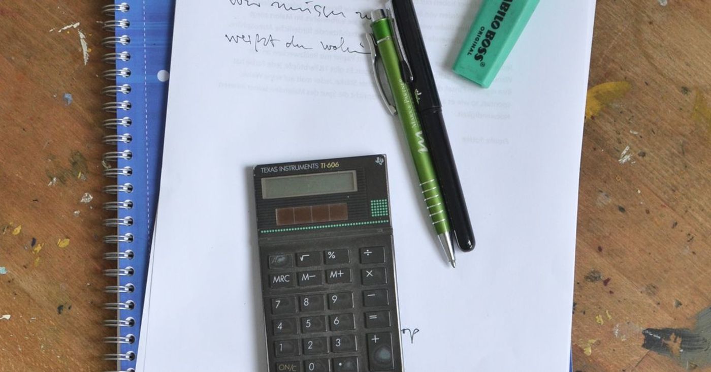 A notebook, paper notes, calculator and pens on a wooden desk.