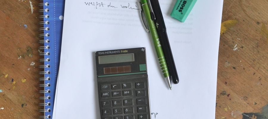 A notebook, paper notes, calculator and pens on a wooden desk.